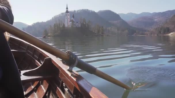 Primer plano cámara lenta de remo remo en un barco en el lago Bled con isla Bled y la iglesia detrás de ella y un reflejo en el lago, con la luz del sol en el barco — Vídeos de Stock