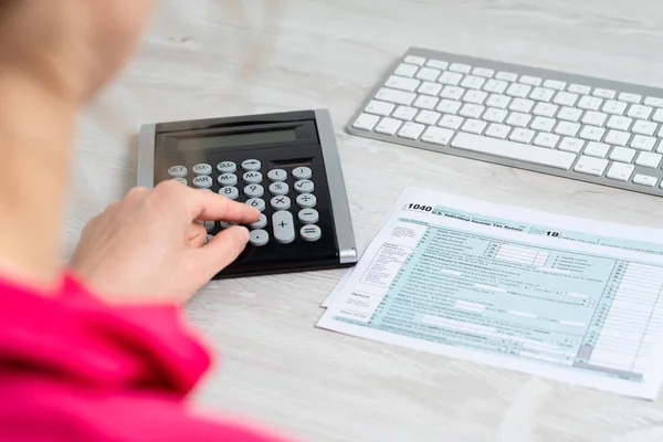 Na zdjęciu ramię kobiety obliczania nam formularz podatkowy obok klawiatury komputera i formularz podatkowy 1040. — Zdjęcie stockowe
