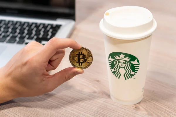 Kryptowährung Bitcoin, gehalten von einer humorvollen Hand neben einer Starbucks-Kaffeetasse. Slowenien 13.02.2019 — Stockfoto