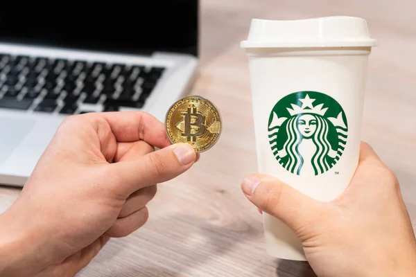 Kryptowährung Bitcoin, gehalten von einer humorvollen Hand neben einer Starbucks-Kaffeetasse. Slowenien 13.02.2019 — Stockfoto