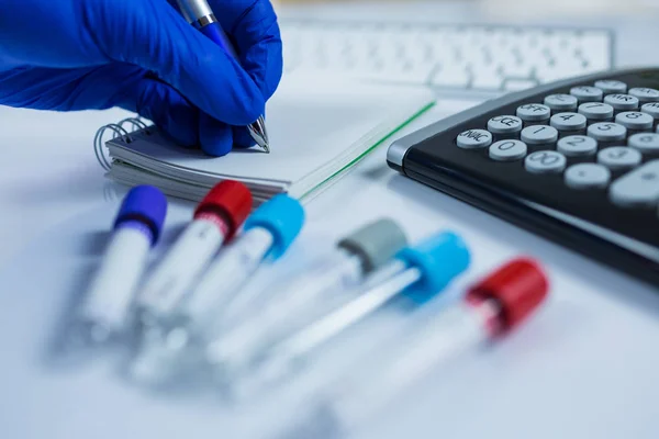 Χέρι γάντια μπλε κάνοντας σημειώσεις δίπλα σε φιάλες για τα δείγματα που χρησιμοποιούνται σε νοσοκομεία ή φάρμακο για τα δείγματα αίματος σε ένα εργαστήριο λευκό φόντο — Φωτογραφία Αρχείου