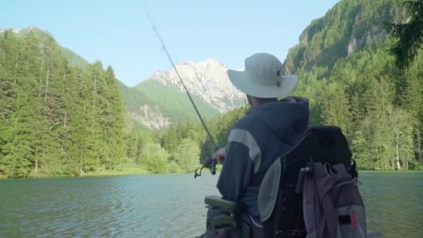 Atrás das costas câmera lenta de pescador deficiente em uma pesca cadeira de rodas elétrica em belo lago perto de floresta e montanha nas costas, durante o dia no verão — Vídeo de Stock