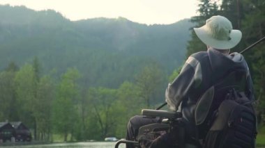 Bir elektrikli tekerlekli sandalye de engelli balıkçı yavaş hareket arka orman ve dağ yakınındaki güzel gölde balıkçılık, gün batımında, yaz