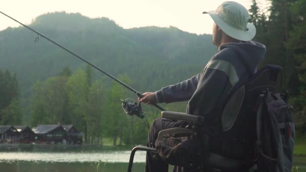 Mouvement lent du pêcheur handicapé dans un fauteuil roulant électrique pêche dans un beau lac près de la forêt et de la montagne à l'arrière, au coucher du soleil, en été — Video