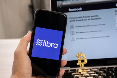 18 Haziran 2019, Ljubljana Slovenya - Üzerinde Terazi logosu olan bir akıllı telefonu tutan el, bilgisayar dizüstü bilgisayarında açılan Facebook web sitesinin yanında