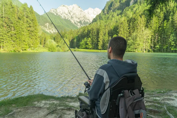 Junger Mann im Rollstuhl angelt an einem sonnigen Tag am schönen See, mit Bergen im Rücken — Stockfoto