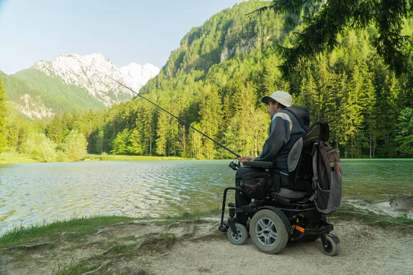 Joven en silla de ruedas pescando en el hermoso lago en un día soleado, con montañas en la parte posterior —  Fotos de Stock