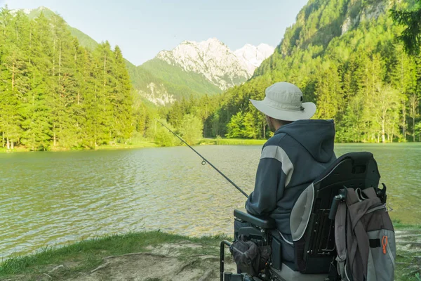 Junger Mann im Rollstuhl angelt an einem sonnigen Tag am schönen See, mit Bergen im Rücken — Stockfoto