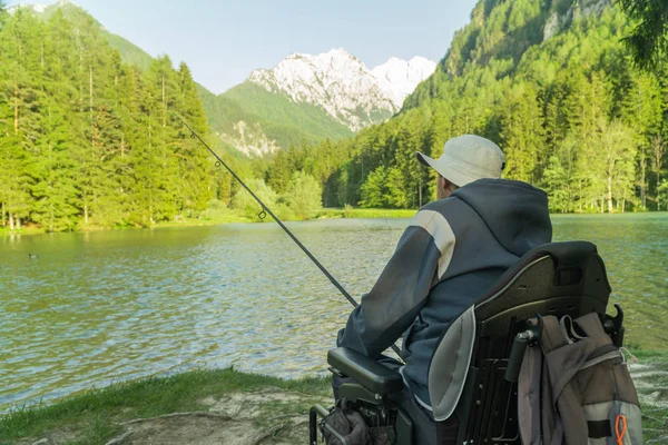 Fiatal férfi tolószékben horgászik a gyönyörű tónál egy napsütéses napon, hátul hegyekkel — Stock Fotó
