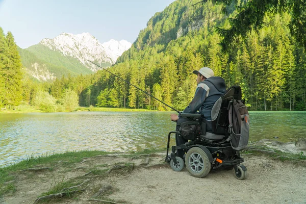 Joven en silla de ruedas pescando en el hermoso lago en un día soleado, con montañas en la parte posterior —  Fotos de Stock