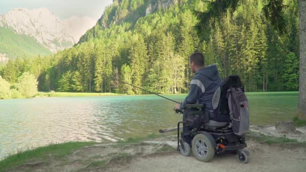 Mouvement lent du pêcheur handicapé dans un fauteuil roulant électrique pêche dans un beau lac près de la forêt et la montagne à l'arrière — Video
