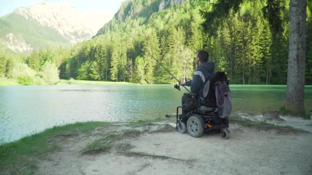 4k-resolutie van gehandicapte visser in een elektrische rolstoel vissen in een prachtig meer in de buurt van bos en berg in de rug — Stockvideo