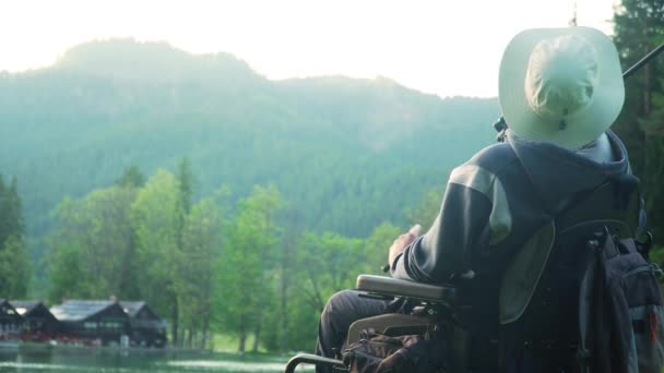 Resolución 4k de pescador discapacitado en una silla de ruedas eléctrica que pesca en hermoso lago cerca del bosque y la montaña en la parte posterior, en la puesta del sol, verano — Vídeo de stock