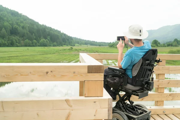 Hombre en silla de ruedas eléctrica con cámara de teléfono inteligente cerca del lago en la naturaleza —  Fotos de Stock