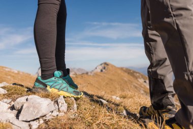 İki yürüyüşçü açık mavi gökyüzünün altında nefes kesici bir dağ manzarasıyla çevrili kayalık arazide duruyor..