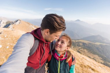 Genç çiftler birbirlerine gülümserler, doğadaki yürüyüş maceraları sırasında büyüleyici dağ manzarasını kucaklarlar..