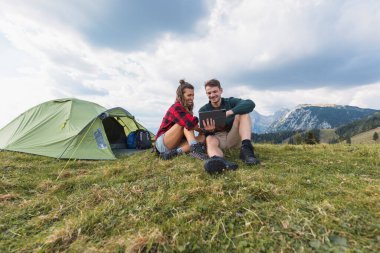 Mutlu genç çift kamp yapıyor, çadırlarının dışında bir tablet üzerinde bir şey izliyor, doğayla çevrili neşeli bir anı paylaşıyorlar..