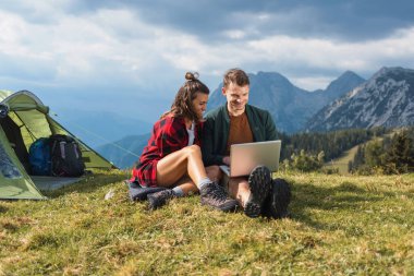 Çimenli bir tepede oturan bir çift, etrafları çarpıcı dağ manzarası ve çadırlarıyla çevriliyken dizüstü bilgisayarla çalışıyor..