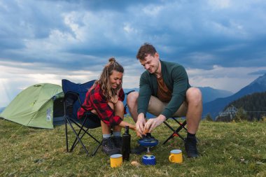 Genç bir çift, kaygısız bir kamp sabahının tadını çıkarıyor çadırlarının önünde eğleniyor, gaz ocağında krep yapıyor..