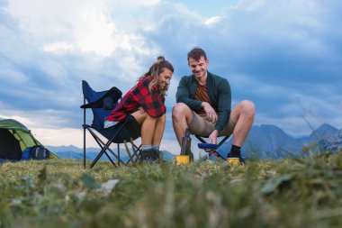 Bir çift kamp sandalyesinde oturur, gülerek açık havada, aydınlık bir günde, dağ manzarası arasında yemek hazırlarlar..