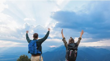İki yürüyüşçü bir dağın zirvesinde sevinir, kolları zaferle yükselir, günbatımında geniş vadilere ve uzak sıradağlara bakar..