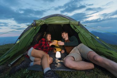 İki insan çadırda dinleniyor, içkilerini paylaşıyor ve bulutlu bir gökyüzünün altında manzaralı dağlarla çevrili huzurlu bir akşamın tadını çıkarıyor..