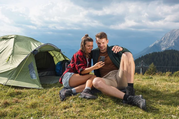 Romantik kamp çifti akıllı telefonlarıyla dışarıda anları yakalıyorlar, manzaralı bir dağ manzarasında çadırın yanında oturuyorlar..