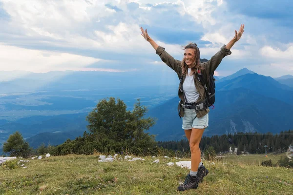 Mutlu bir yürüyüşçü, kollarını bir dağın zirvesinde kaldırır. Etrafı geniş manzaralarla ve arka planda güneş batarken uzak dağlarla çevrilidir..