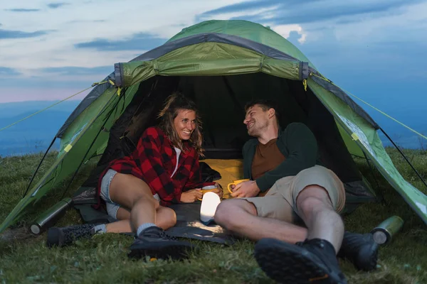 Bir çift, dağdaki yeşil bir çadırda dinleniyor sıcak içeceklerin ve arkadaşlığın tadını çıkarırken rahat bir anı paylaşıyor..