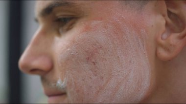 Genç bir adam, sabah rutini sırasında cilt bakımına odaklanarak iyi aydınlatılmış bir banyoda yüzüne krem sürüyor..