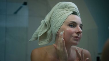 Modern bir banyoda yüzüne nemlendirici süren bir kadın havluya sarılıp cilt bakımını yapıyor..