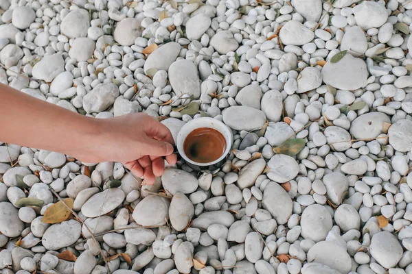 Hand pick up a black cup of hot coffee on white rock in garden floor. Background and texture of white pebbles stone. Take a break from work.