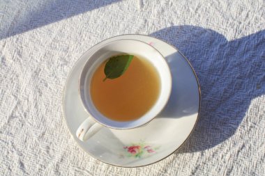 Nane yaprağıyla sıcak çay, beyaz lüks bardakta, güneş ışığı altında ahşap tepside. Sağlık ve bitkiler için sabah kahvaltısı.