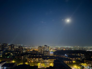 Şehir geceleri gökyüzünde parlayan bir dolunayla aydınlanıyor. Ay gökyüzünün ortasında konumlanmış, şehrin üzerine yumuşak bir parıltı yayıyor.