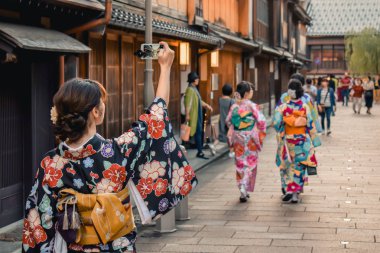 Japanese girl in kimono taking a photo of a traditional street with wooden houses on her cell phone in Kanazawa Japan clipart
