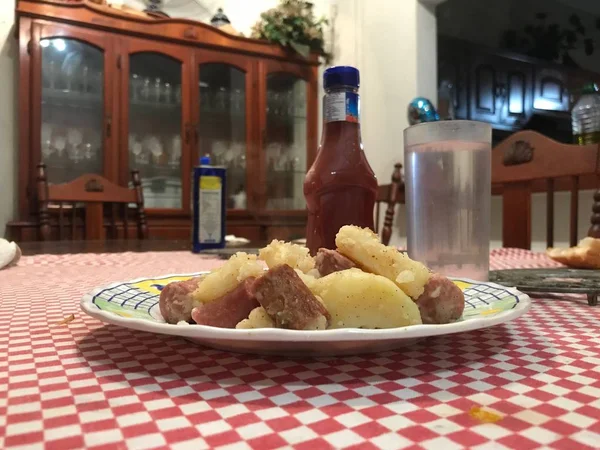 Placa de comida preparada a base de salchichas fritas y patatas cocidas al frente y vaso de agua y botella de salsa de tomate al fondo . — Foto de Stock