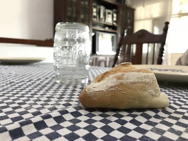 Pezzo di pane davanti con piatto di porcellana e vetro sullo sfondo su una tovaglia con quadrati blu — Foto Stock