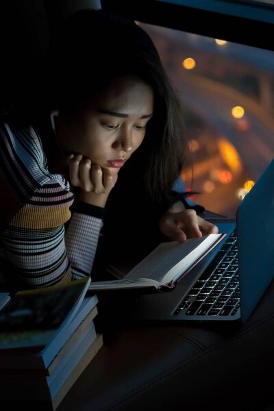 Beautiful young Asian girl reading book and working with a laptop connecting to internet via computer