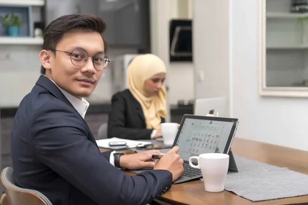 Ásia malaio homem trabalho no casa com laptop com colega no fundo — Fotografia de Stock