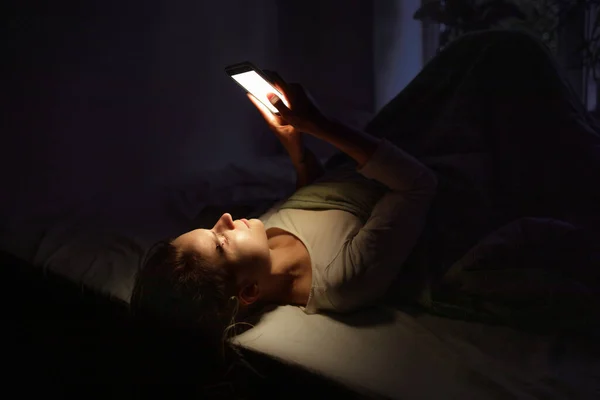 Genç bir kız yatakta yatar ve akıllı telefonuna bakar.