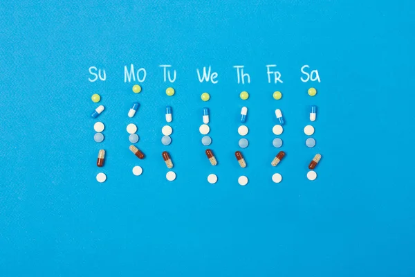 Weekly plan or schedule of medication in pills and capsules with the names of the days of the week on the blue background. Top view.