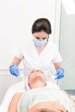 Kozmetik salonunun genç bayan müşterisi özel cihazlarla mikroakım prosedürü uyguluyor, yakın plan. Güzellik uzmanı yüz işlemleri için elektriksel impulslar kullanıyor. Cilt bakımı ürünleri ve güzellik salonu veya klinik kavramları.