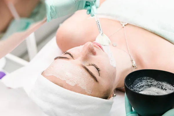 The young female client of cosmetic salon having a cleansing facial mask. The procedure of applying a peeling mask to the face. Concepts of skin care and beauty salon or clinic.