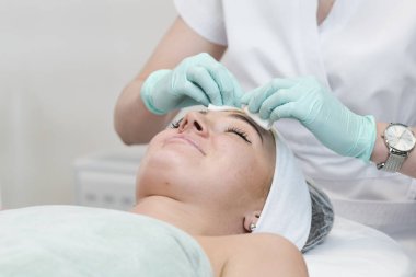 Kozmetik salonunun genç bayan müşterisi yüz temizleme ameliyatı oluyor. Doktor kozmetik uzmanı deriyi süngerle temizliyor. Cilt bakımı ve güzellik salonu veya klinik kavramları.