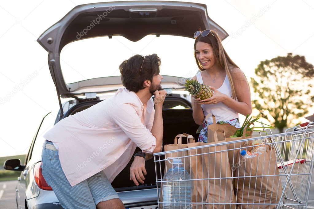 A happy couple puts groceries in his car in front of a supermarket.