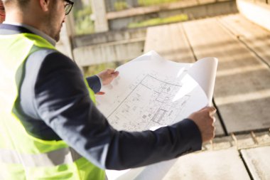 Mimar inşaat alanında, elinde bir inşaat projesi tutuyor..