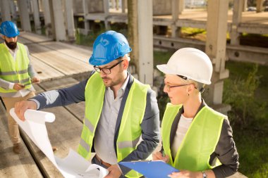 Mimar inşaat alanında, elinde bir inşaat projesi tutuyor..