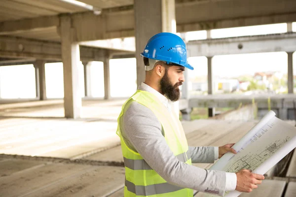 A young man with a beard looks at the construction plans, it is on a building construction site.