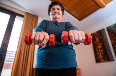 Siyah kısa saçlı yaşlı bir bayanın elinde bulunan küçük kırmızı ağırlıklara odaklanın. Evinde fiziksel durumu kontrol altına alıyor..