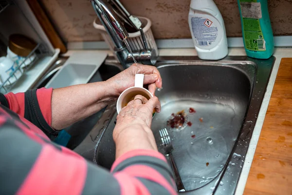 An elderly senior lady stands in the kitchen and washes the dishes by hand. In his hands he washes a cup under running water.